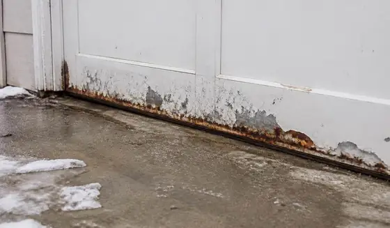Rust and peeling paint along the bottom panel of a white steel garage door in winter